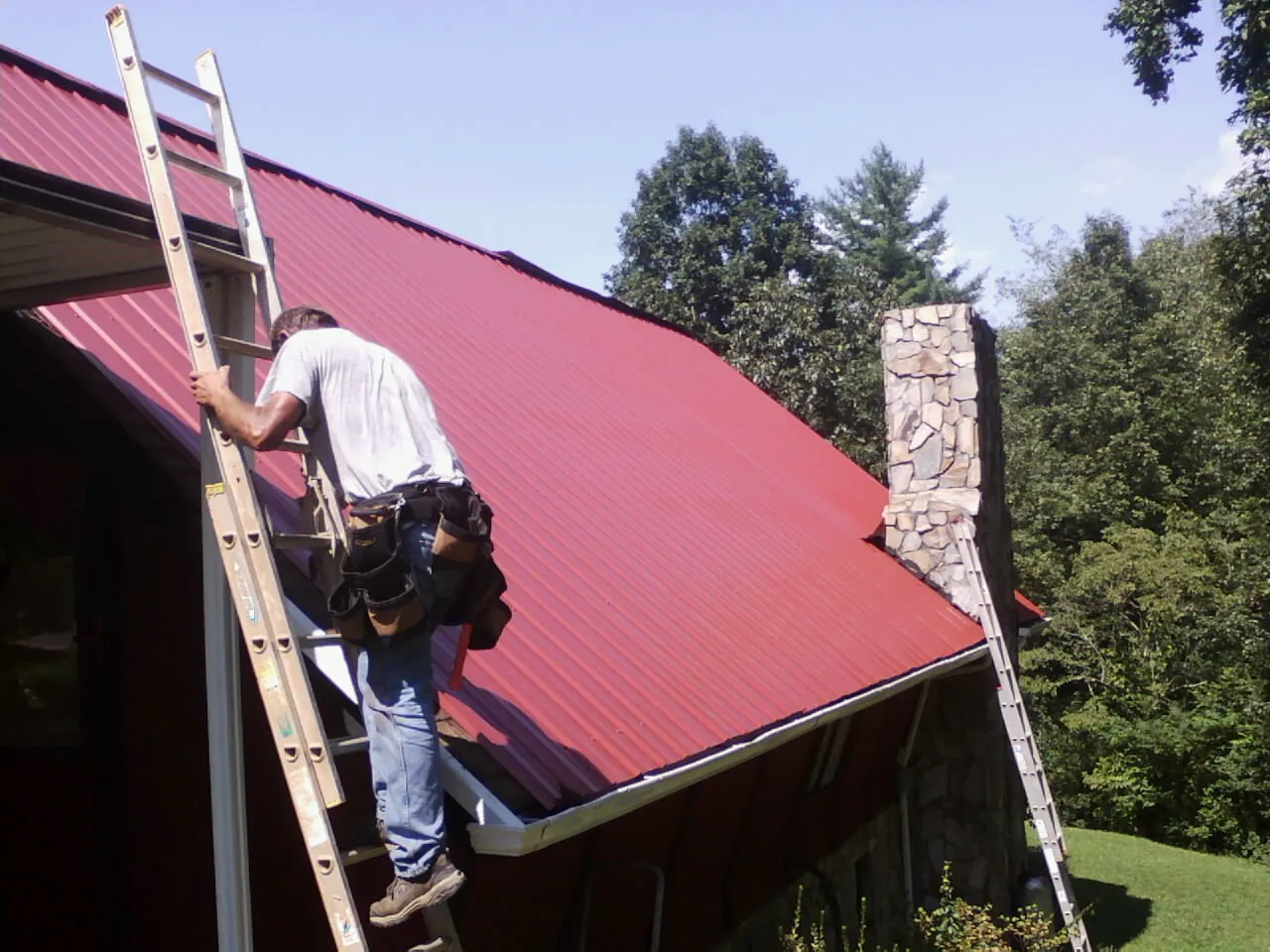 Roofer on a ladder beside a red metal roof and stone chimney — Soffit & Fascia Repair by Roofing Brothers in Davidson